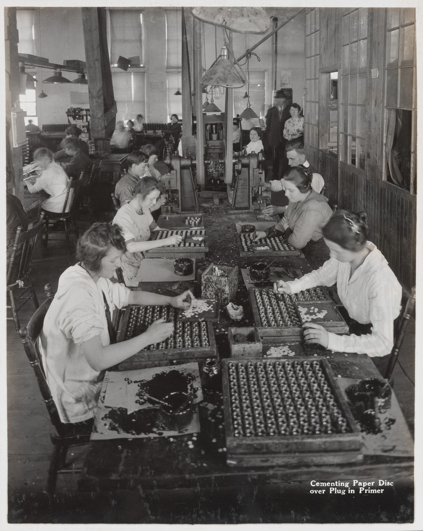 Interior view of workers cementing paper disc over plug in primer at the Energite Explosives Plant No. 3, the Shell Loading Plant, Renfrew, Ontario, Canada