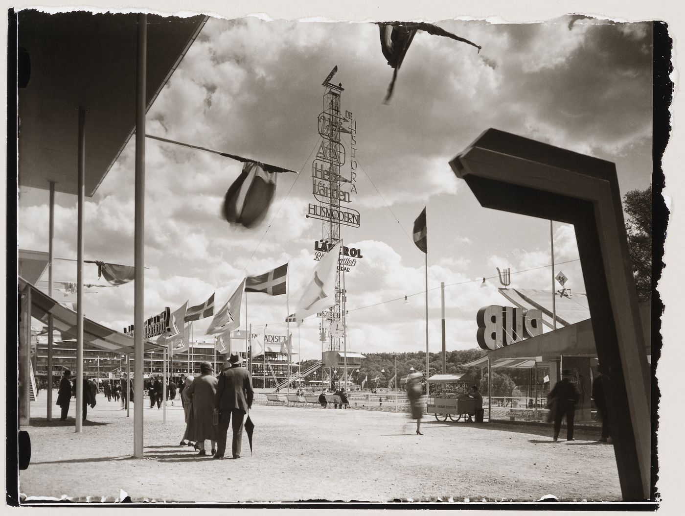 Exterior view of the advertising mast and Paradise Restaurant at the Stockholm Exhibition of 1930 showing a walkway and flagpoles, Stockholm
