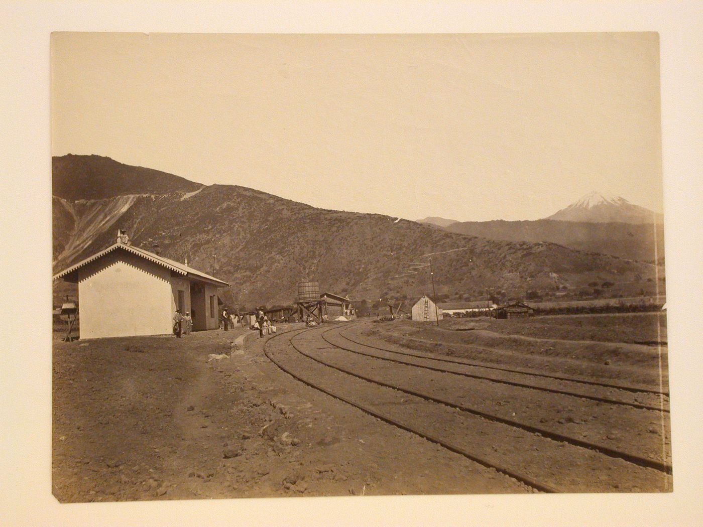 View of the Maltrata railway station showing men and a water tower [?] with mountains and the Pico de Orizaba Volcano in the background, Mexico