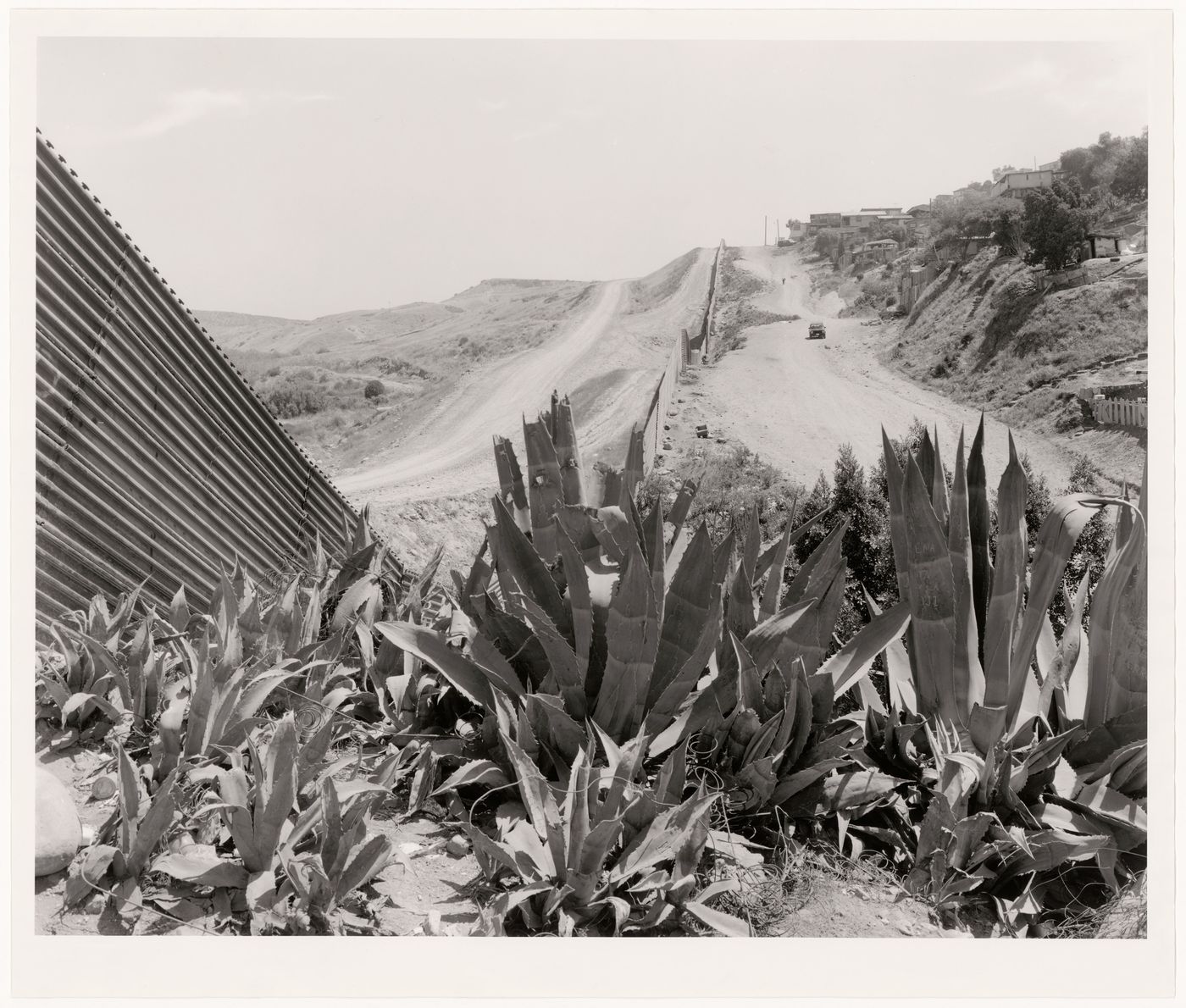 Partial view of the United States-Mexico border fence with plants in the foreground, San Diego County, California, United States, and Colonia Libertad, Tijuana, Baja California, Mexico