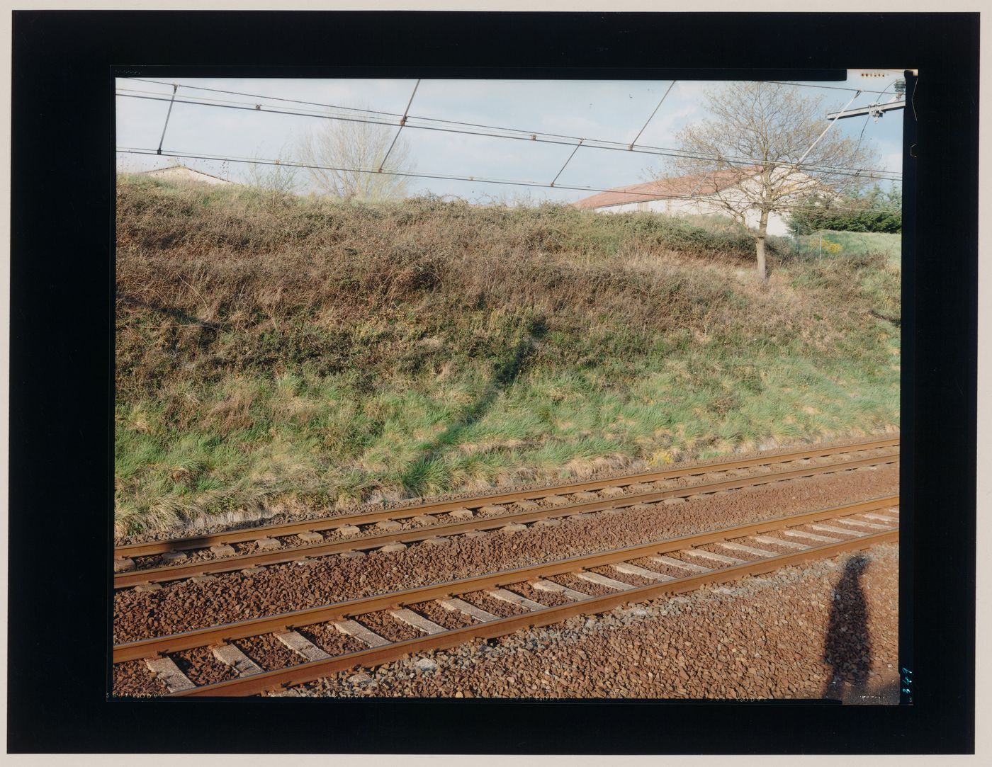 View of tracks and overhead wires for an electric railway, a grassy slope and a person's cast shadow, Orthez, France (from the series "In between cities")