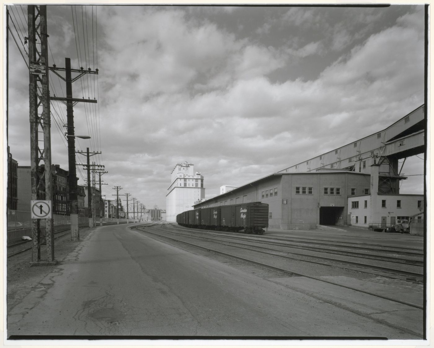 View of Shed No. 11 surmounted by a conveyor gallery with railway cars on the right and Grain Elevator No. 2 (now demolished) in the background, rue de la Commune, Port of Montréal, Québec