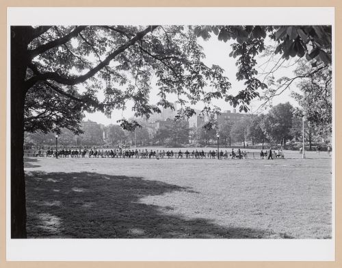 View of King Park field showing a long bench and people sitting with buildings in the background, Long Island