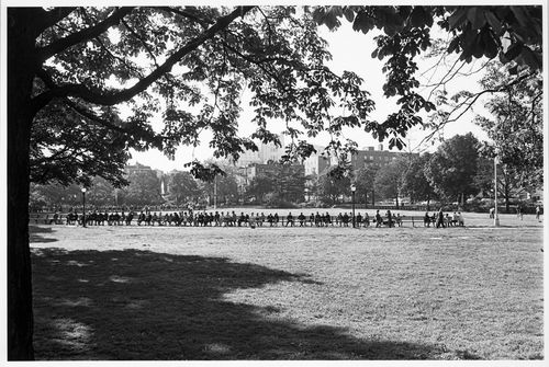 View of King Park field showing a long bench and people sitting with buildings in the background, Long Island