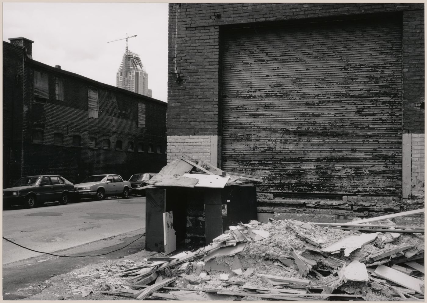 Field Work in Montreal: View of a wooden shack-like structure, a brick wall, an overhead door, buildings and parked automobiles showing Le 1000 de La Gauchetière under construction in the background, Montréal, Québec