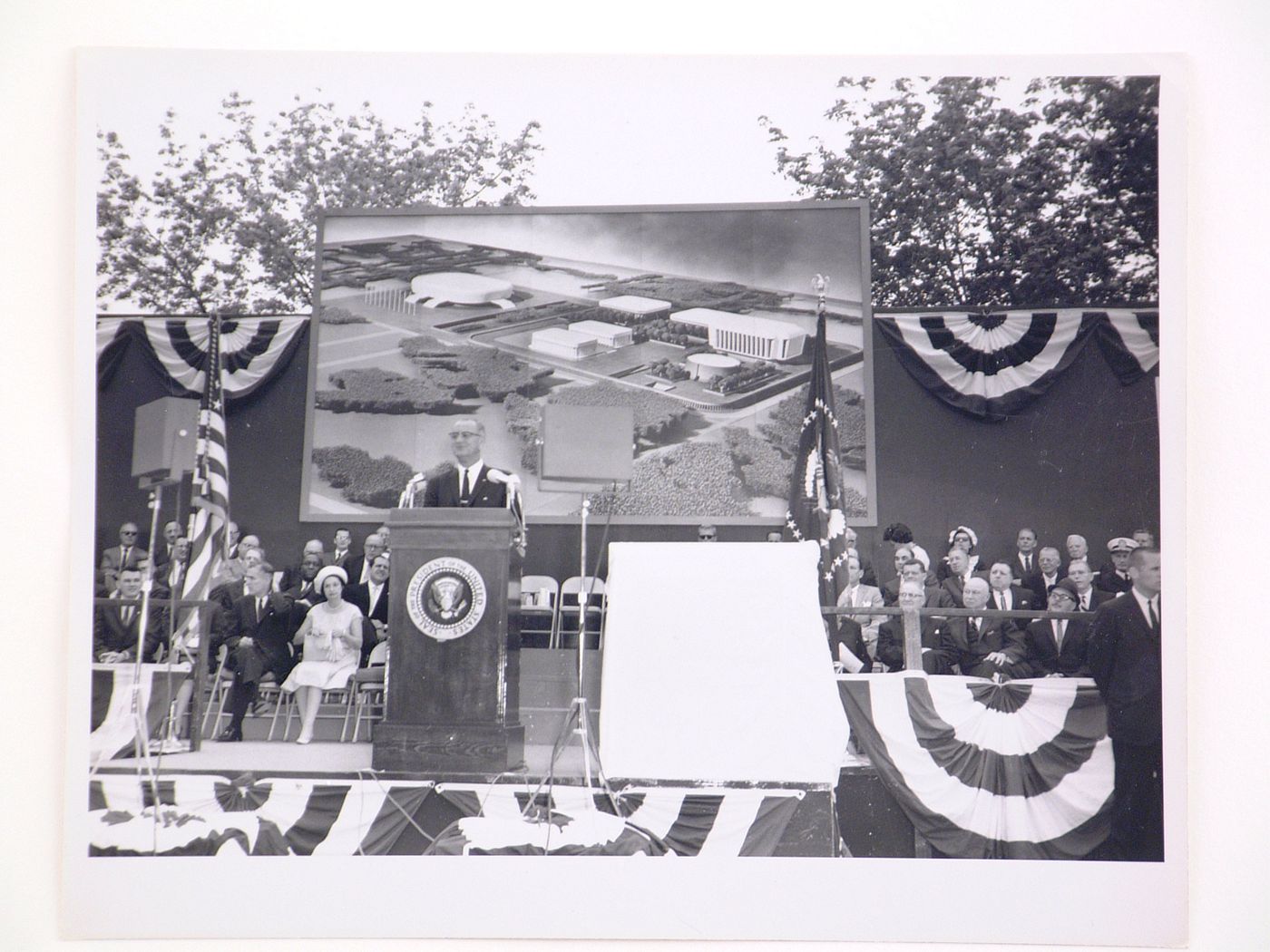 View of the dedication ceremony of the John F. Kennedy Educational, Civic and Cultural Center showing a partial view of the platform with people, posters of President Lyndon B. Johnson and a photograph of the model of the building, Mineola, New York