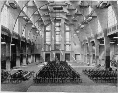 Interior view of a building hall, possibly a church, showing arches, windows and aligned chairs