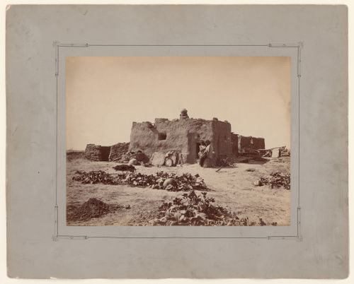 View of Zuni adobe house showing a woman and child seated at entrance and squash planted in the foreground, probably Arizona or New Mexico, United States