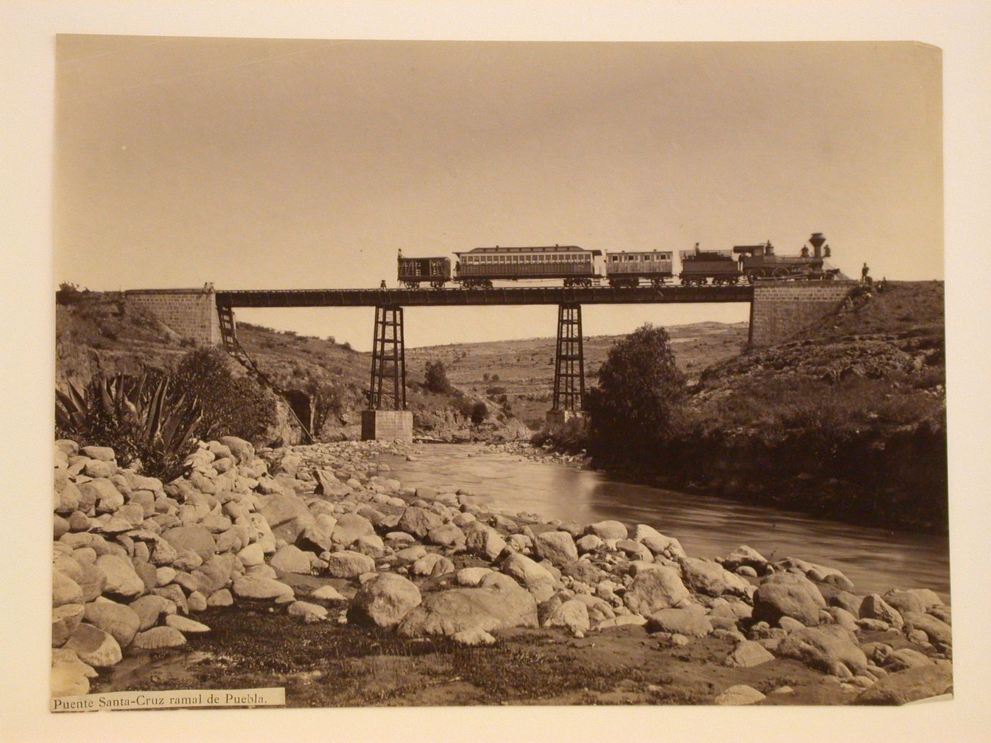 View of a train and people on the Santa-Cruz railroad bridge showing boulders and a river in the foreground, Puebla [?], Mexico