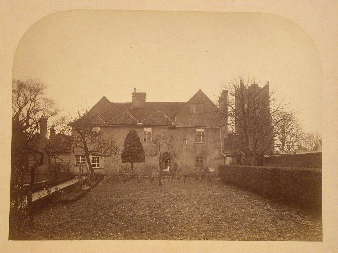 View across a backyard towards a house showing people and trees with what is probably the church tower of St. Mary the Virgin in the distant right, Ardleigh, Essex, England