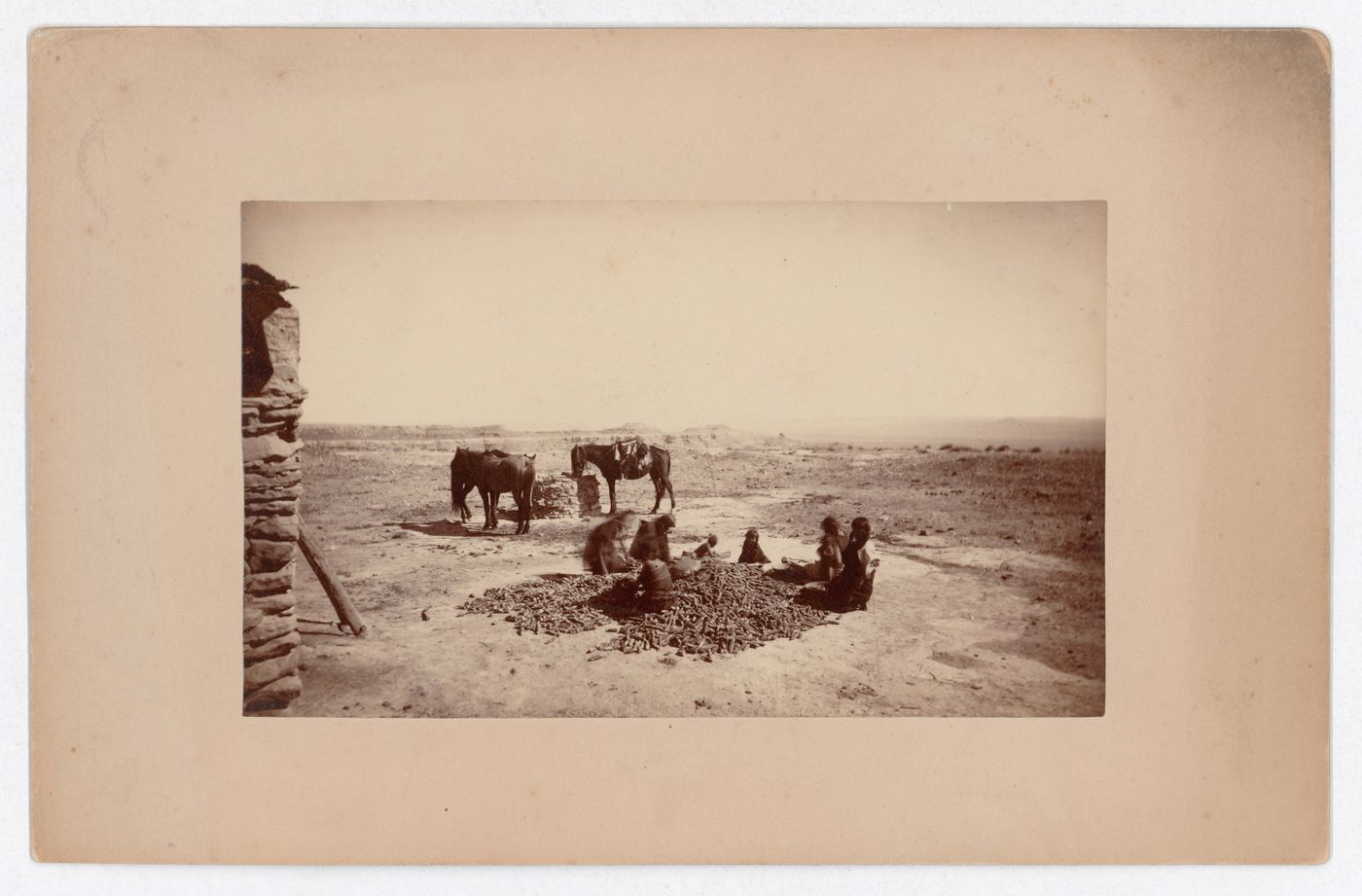 View of children and adults processing corn, with horses around a well in the background, Southwestern United States
