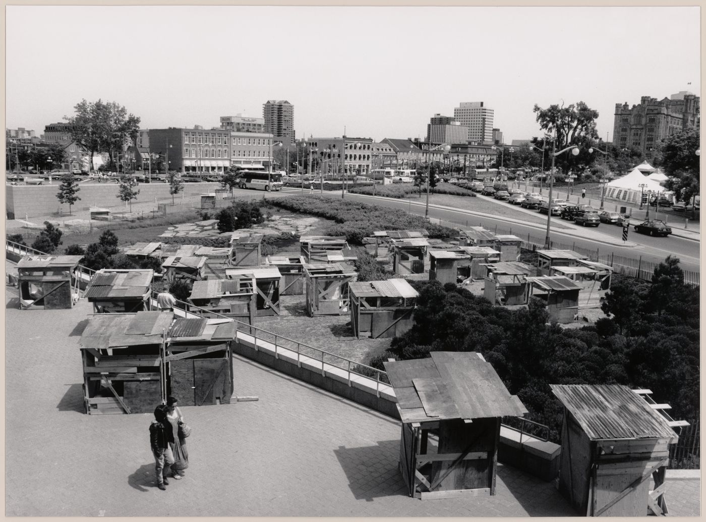 View of the installation "Favela in Ottawa", comprising wooden shack-like structures in the Taiga Garden of the National Gallery of Canada, showing buildings and streets in the background, Ottawa, Ontario