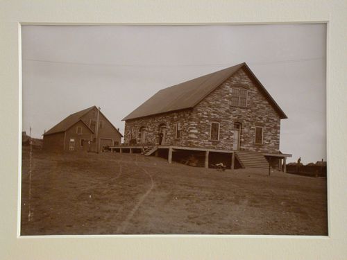 View of stone and wooden mine buildings, one figure at doorway, another sitting on containers