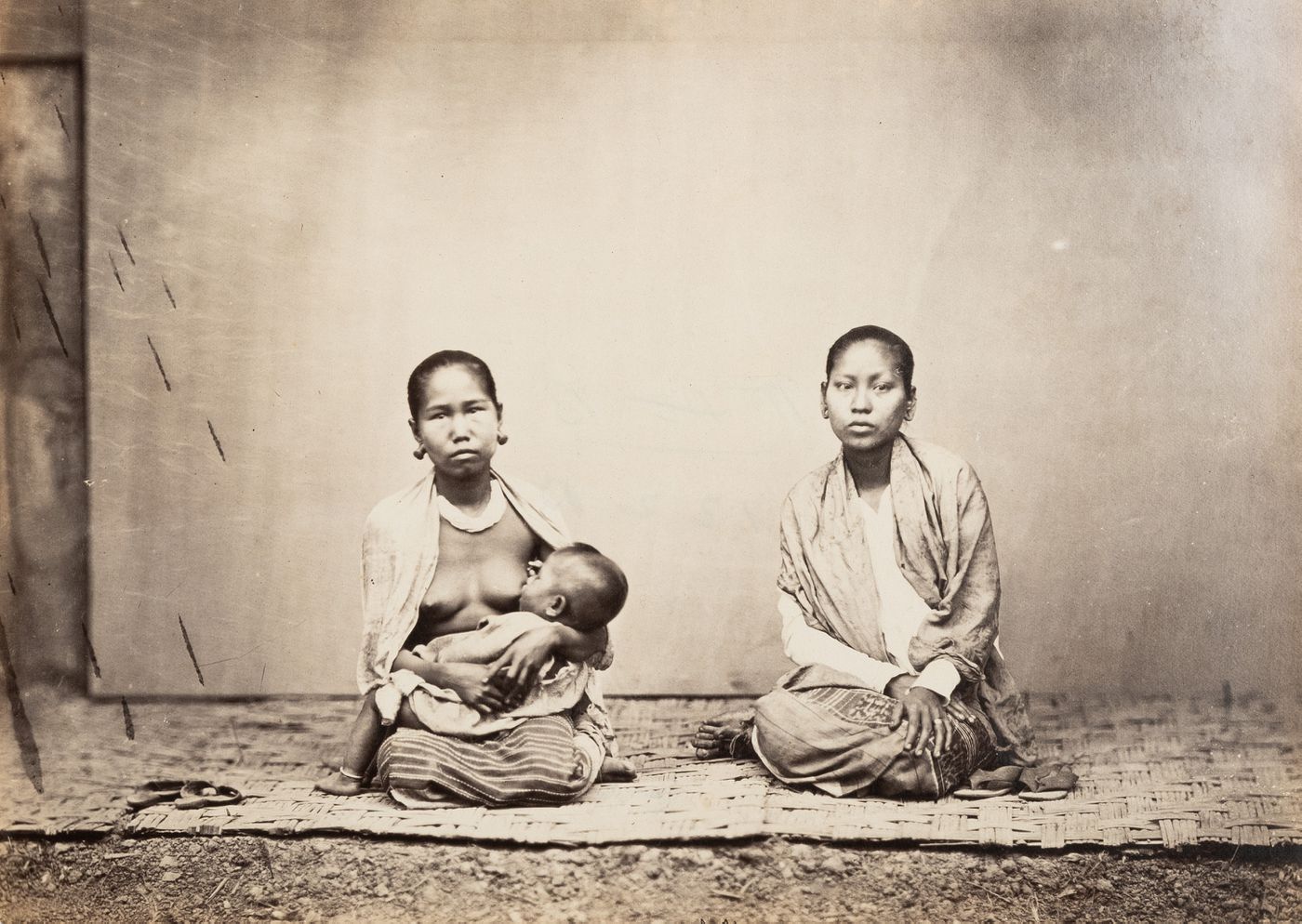 View of two seated Burmese women showing one breast-feeding a child, Burma (now Myanmar)
