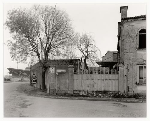 View of buildings and a gate with a ship's bow in the background, on the corner of Via A. Volta, Marghera, Italy