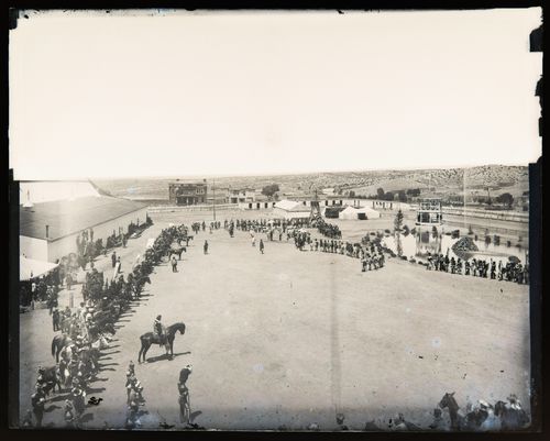 View of a fortified (?) camp where a large gathering of Indigenous people and uniformed men on horseback is taking place, (possibly) Arizona, United States of America
