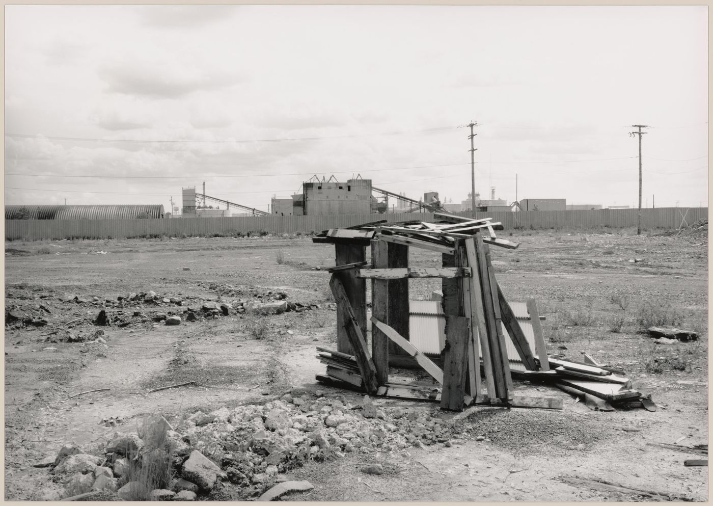 Field Work in Montreal: View of a shack-like structure of wood and corrugated fibreglass, a vacant lot and utility poles showing a metal fence and a processing plant in the background, Montréal, Québec