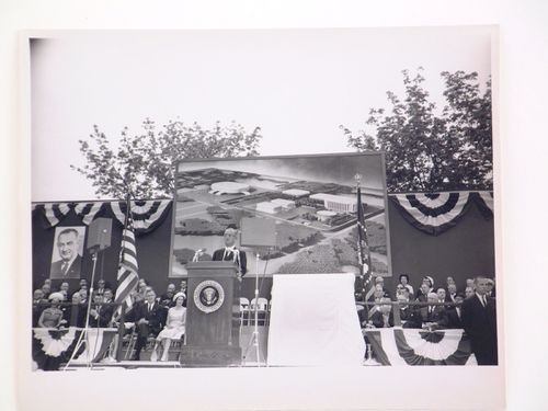 View of the dedication ceremony of the John F. Kennedy Educational, Civic and Cultural Center showing a partial view of the platform with people, posters of President Lyndon B. Johnson and a photograph of the model of the building, Mineola, New York
