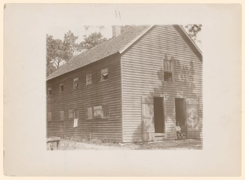 Exterior view of wooden building containing twenty-four rooms occupied by cranberry pickers on Forsythe's Bog, with a small child seated in doorway, near Pemberton, New Jersey, United States