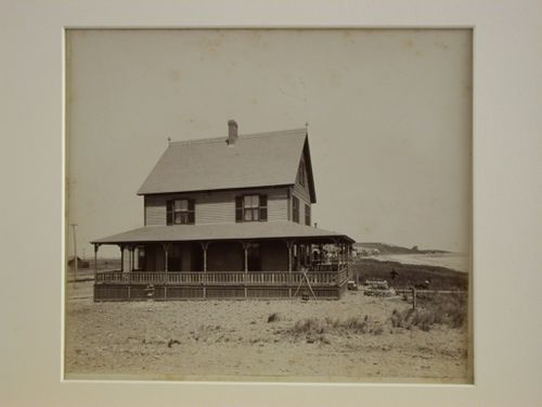 Wooden house with wrap-around porch, near shore: two figures in grass at right