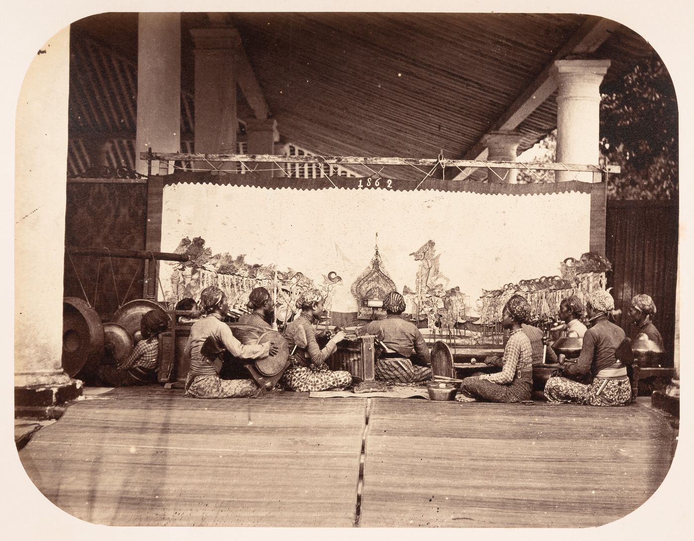 Interior view of a shadow theatre showing the musicians and instruments of a gamelan, shadow puppets and a building, probably in Bandong (now Bandung), Dutch East Indies (now Indonesia)