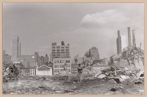View of a demolition site [?] showing a man and fire hose with skyscrapers and smokestacks in the background, New York City, New York