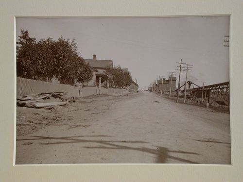 Looking down a street, fence and houses on one side, trestle and wooden mine shaft buildings on the other side