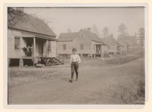 Photograph of Louis Stokes, one of the young workers in the Kosciusko Cotton Mills, walking along street with wooden houses, Kosciusko, Mississippi, United States