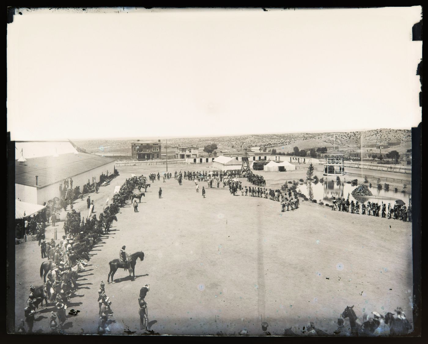 View of a fortified (?) camp where a large gathering of Indigenous people and uniformed men on horseback is taking place, (possibly) Arizona, United States of America