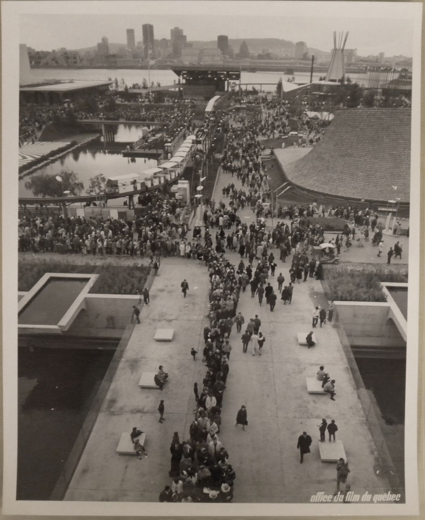 View of a walkway with visitors standing in line near the Pavilion of Western Canada with the Atlantic Provinces', the Indians of Canada Pavilions and Montréal in background, Expo 67, Montréal, Québec