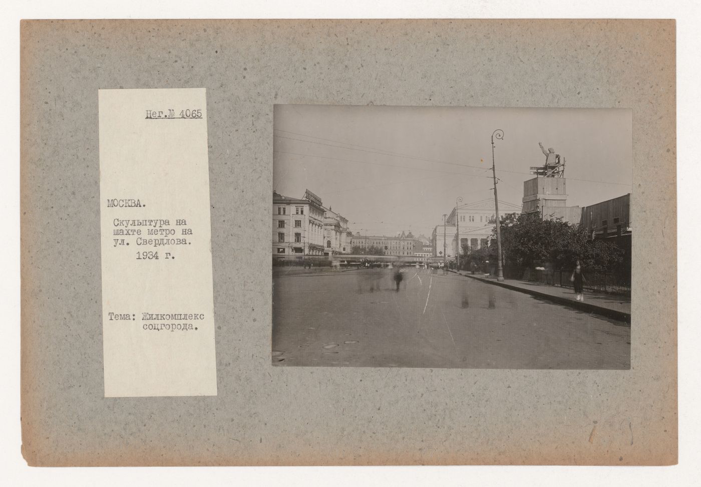 View of Sverdlova Square (now Teatral'naia Square) showing the subway station under construction surmounted by a large wooden figure of a worker and surrounded by hoardings, with the Bol'shoi Theater in the background, Moscow