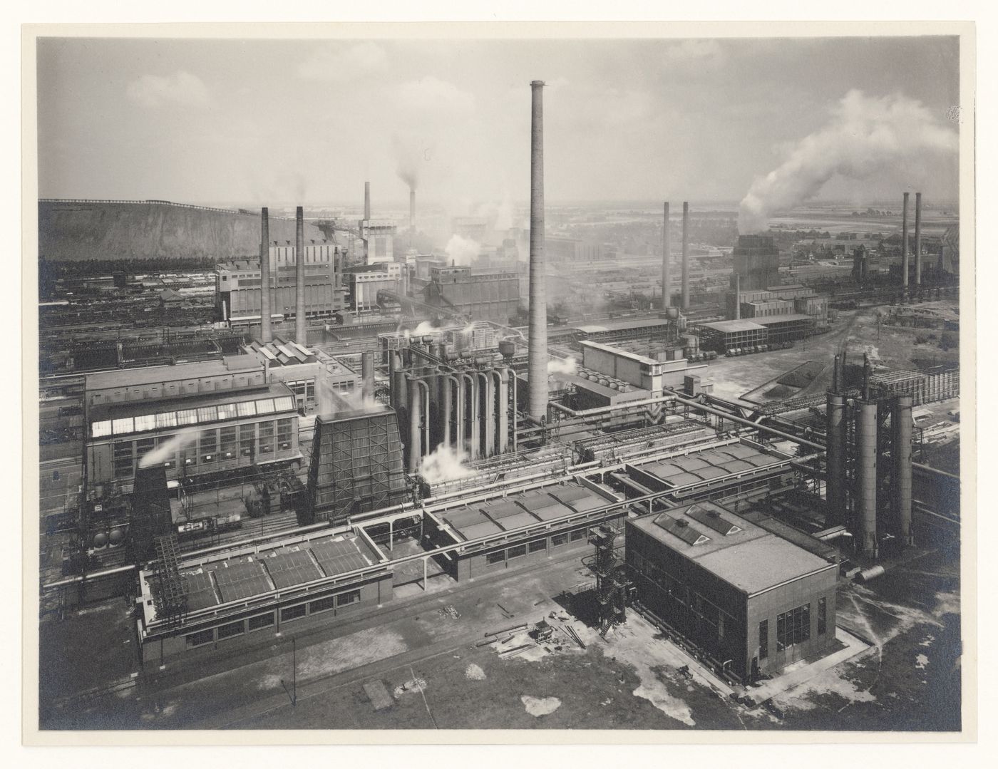 General view of large industrial site, from elevated view point, showing various chimneys, there is a tall one in the center of the composition