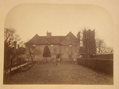 View across a backyard towards a house showing people and trees with what is probably the church tower of St. Mary the Virgin in the distant right, Ardleigh, Essex, England