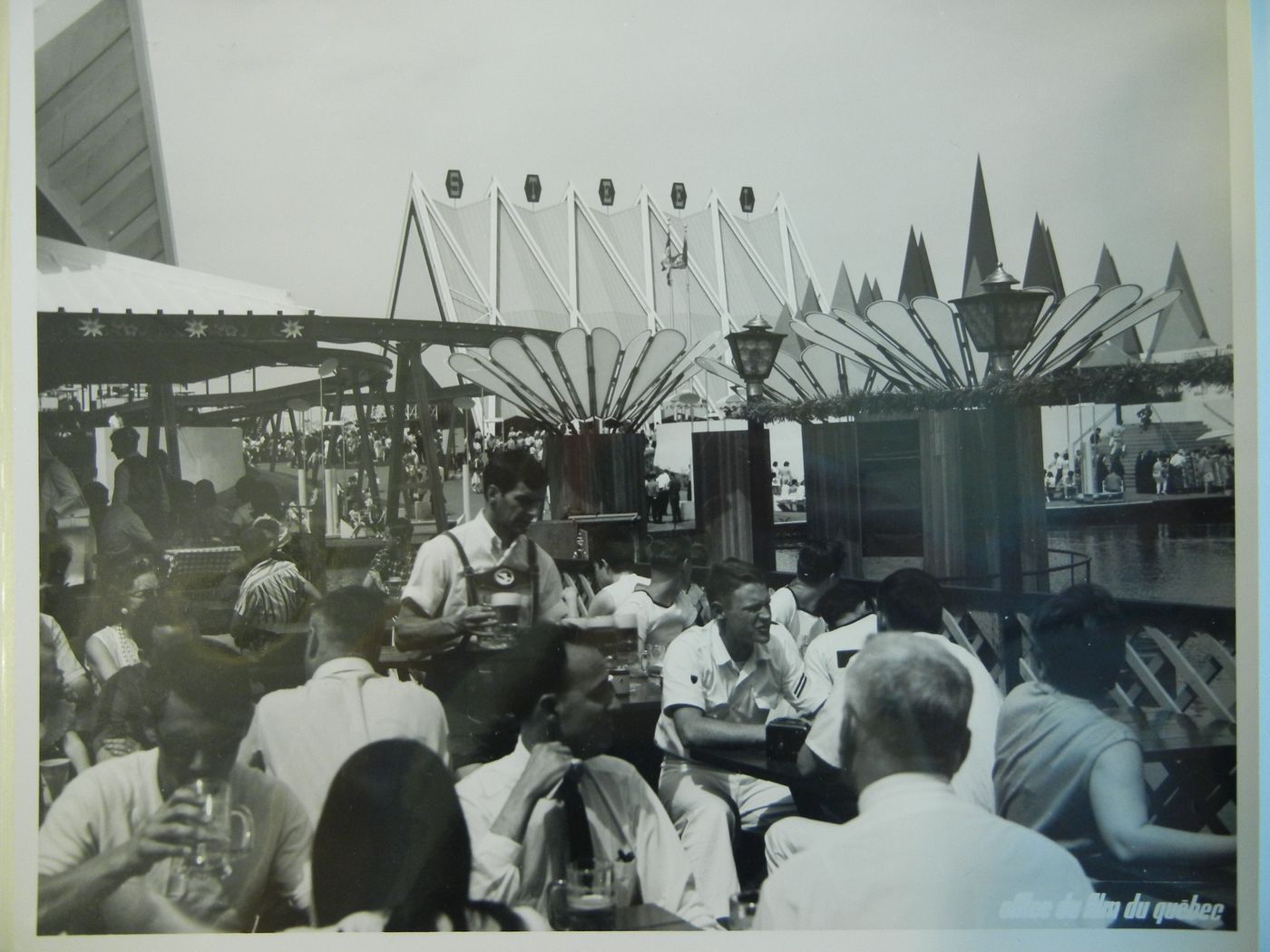 View of seated visitors drinking beer at the Expo-Services C with the Steel Pavilion in background, Expo 67, Montréal, Québec