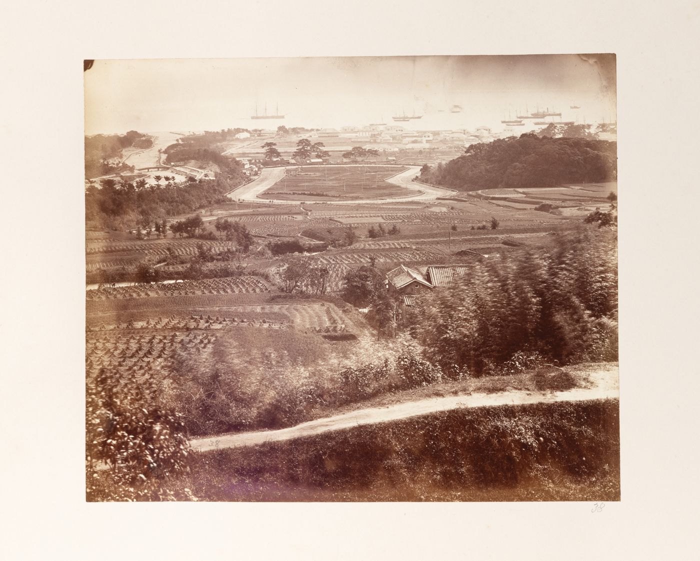 View of a horse racetrack showing farmland and a farm in the foreground and part of the foreign settlement in the background, Kobe, Japan
