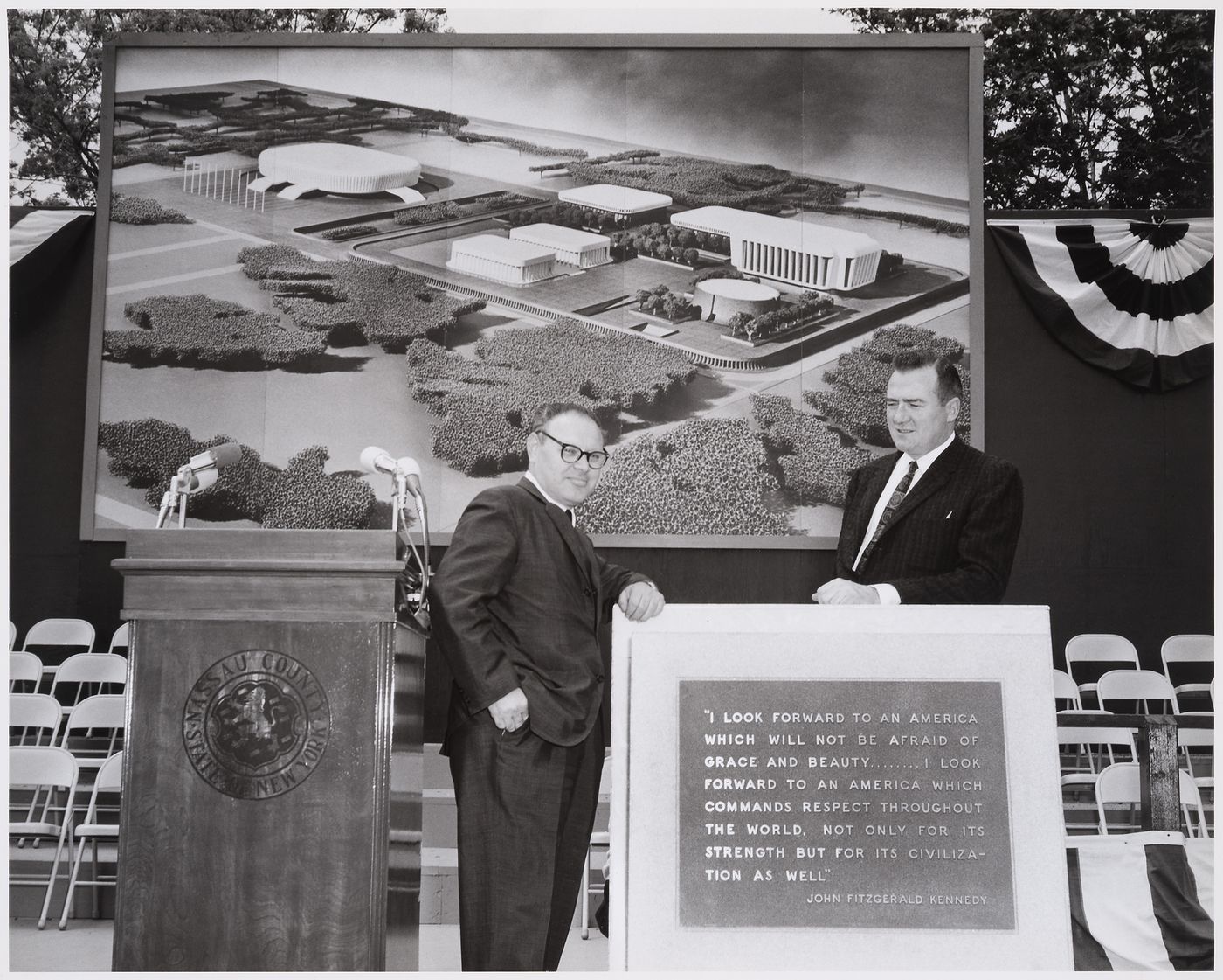 View of the dedication ceremony of the John F. Kennedy Educational, Civic and Cultural Center showing the memorial plaque with a quote by John F. Kennedy, two men and a photograph of the model of the building, Mineola, New York