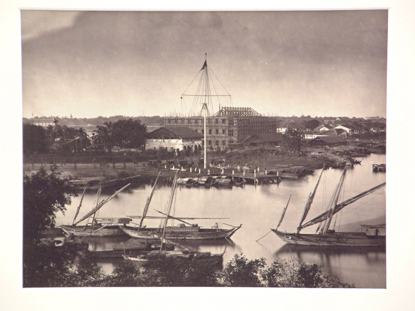 View of the flagtower and junks from across the Saigon River [?] with a building under construction in the background, Saigon (now Ho Chi Minh City), Cochin China (now in Vietnam)