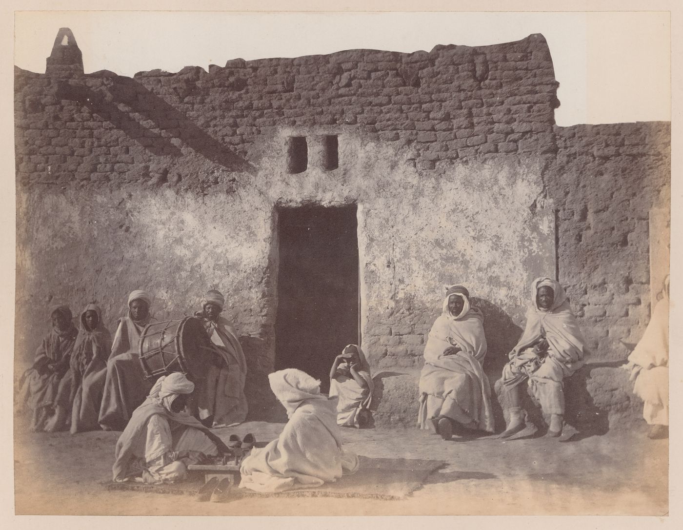 View of people with drum and board game sitting in front of adobe structure, Algiers, Algeria