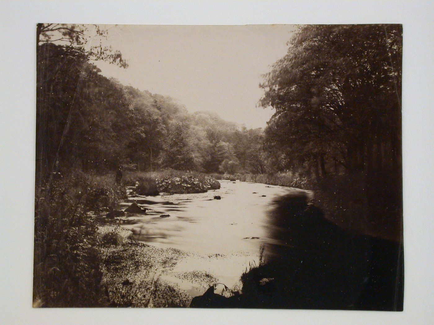 View of a river surrounded by bush and trees with two men on the shore, possibly in the park of Chatsworth House or Haddon Hall, Derbyshire, England