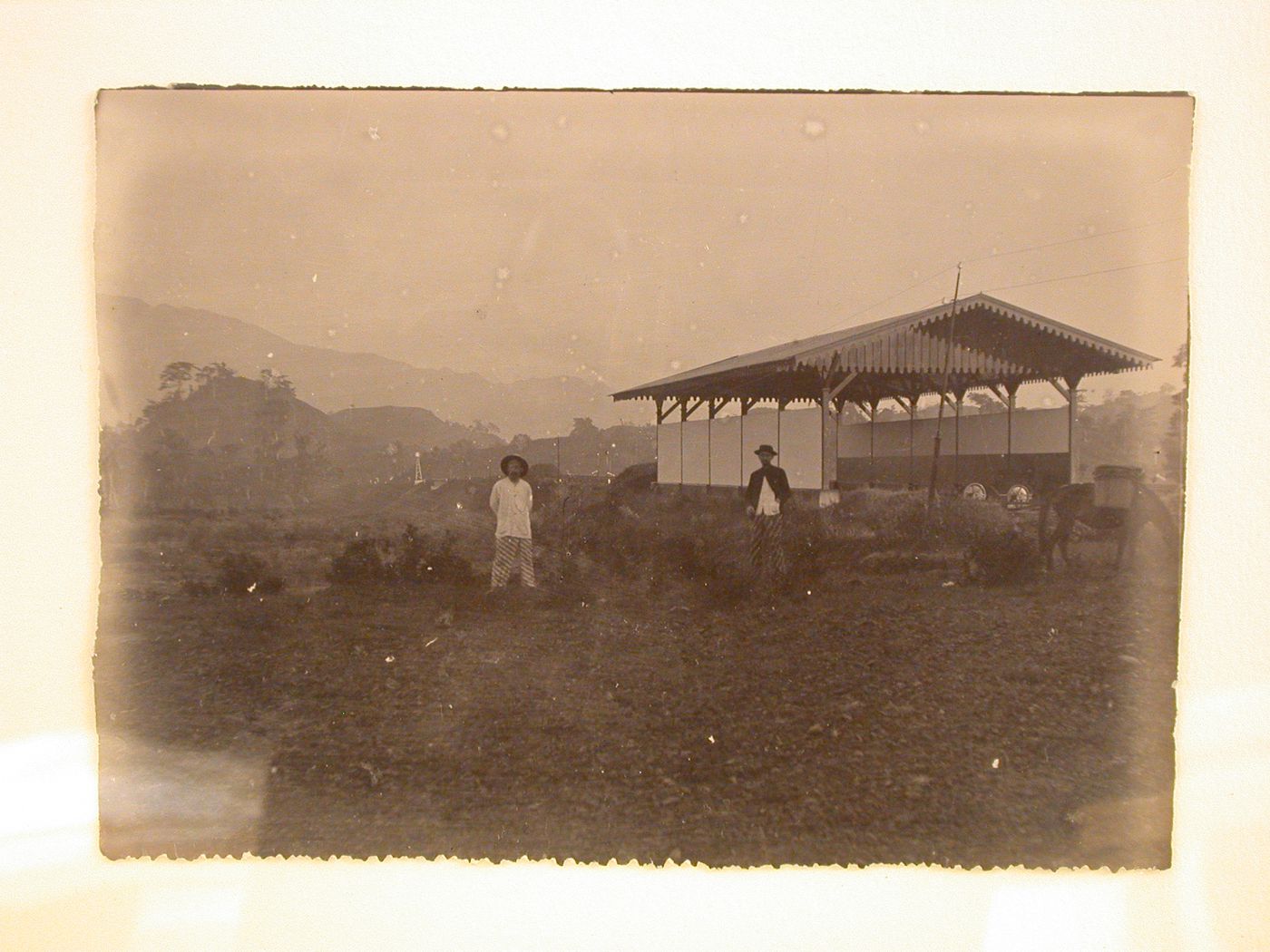 View of two men and a donkey in front of a barn [?] with hills in the distance