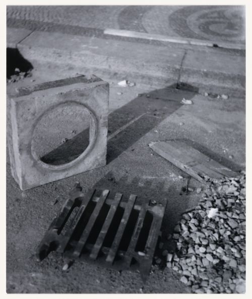 View of a concrete plumbing [?] component, a grate, a wood scrap and gravel showing a mosaic walkway in the background, Berlin, Germany, from the artist book "The Potsdamer Project"