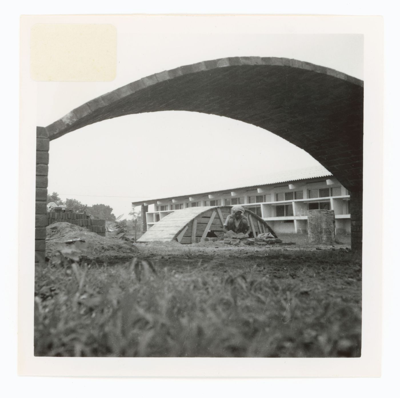 View of arched structures and a man squatting with an unidentified building in background, possibly in Chandigarh, India