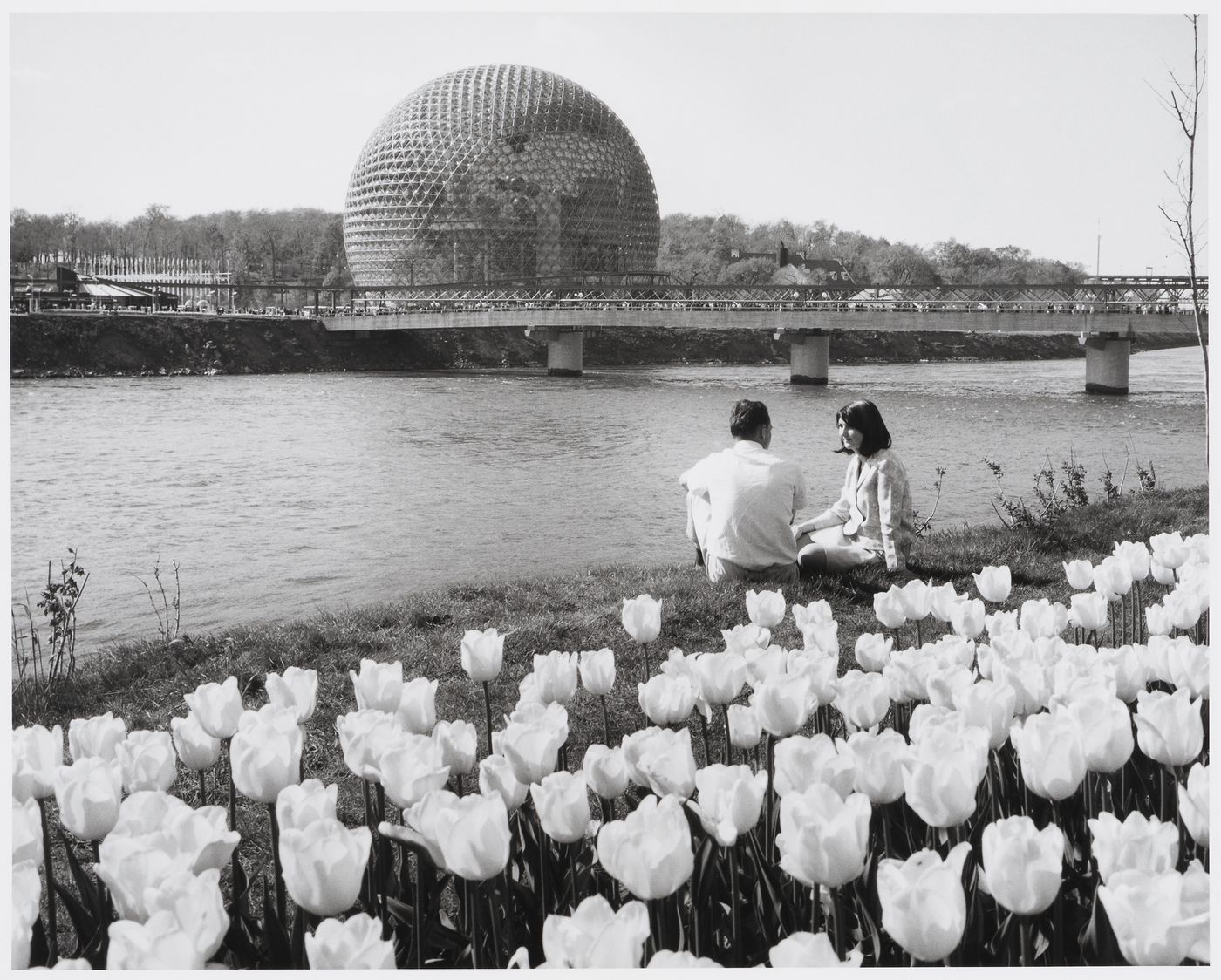 View on the Pavilion of the United States and the Cosmos Walk with a sitting couple surrounded by flowers in foreground, Expo 67, Montréal, Québec