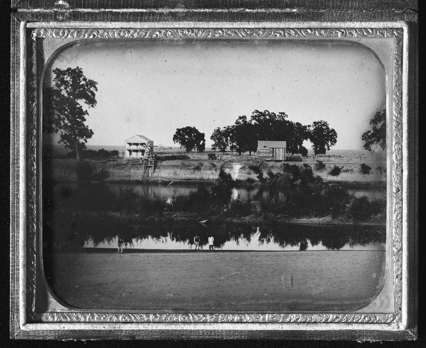 View of balloon-framed farmhouse, windmill on riverside with people on both sides of riverbank, probably United States