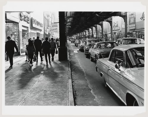View of Jamaica Avenue showing people walking on the sidewalk and cars stopped in a traffic jam under an elevated subway line, Long Island, United States