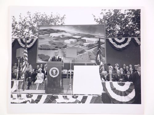 View of the dedication ceremony of the John F. Kennedy Educational, Civic and Cultural Center showing a partial view of the platform with people, posters of President Lyndon B. Johnson and a photograph of the model of the building, Mineola, New York