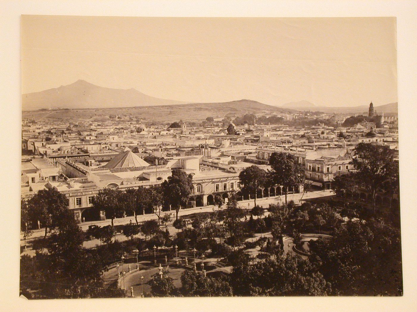 View of the city of Puebla with a public garden in the foreground, hills and volcanoes [?] in the background, and the tower and the principal façade of the Church of San Francisco on the right, Mexico