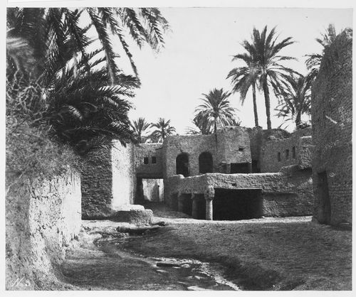 View of stone or sandstone buildings with mostly dried river bed in foreground within an oasis. Buildings look abandoned.