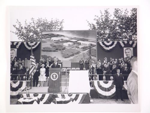 View of the dedication ceremony of the John F. Kennedy Educational, Civic and Cultural Center showing a partial view of the platform with people, posters of President Lyndon B. Johnson and a photograph of the model of the building, Mineola, New York