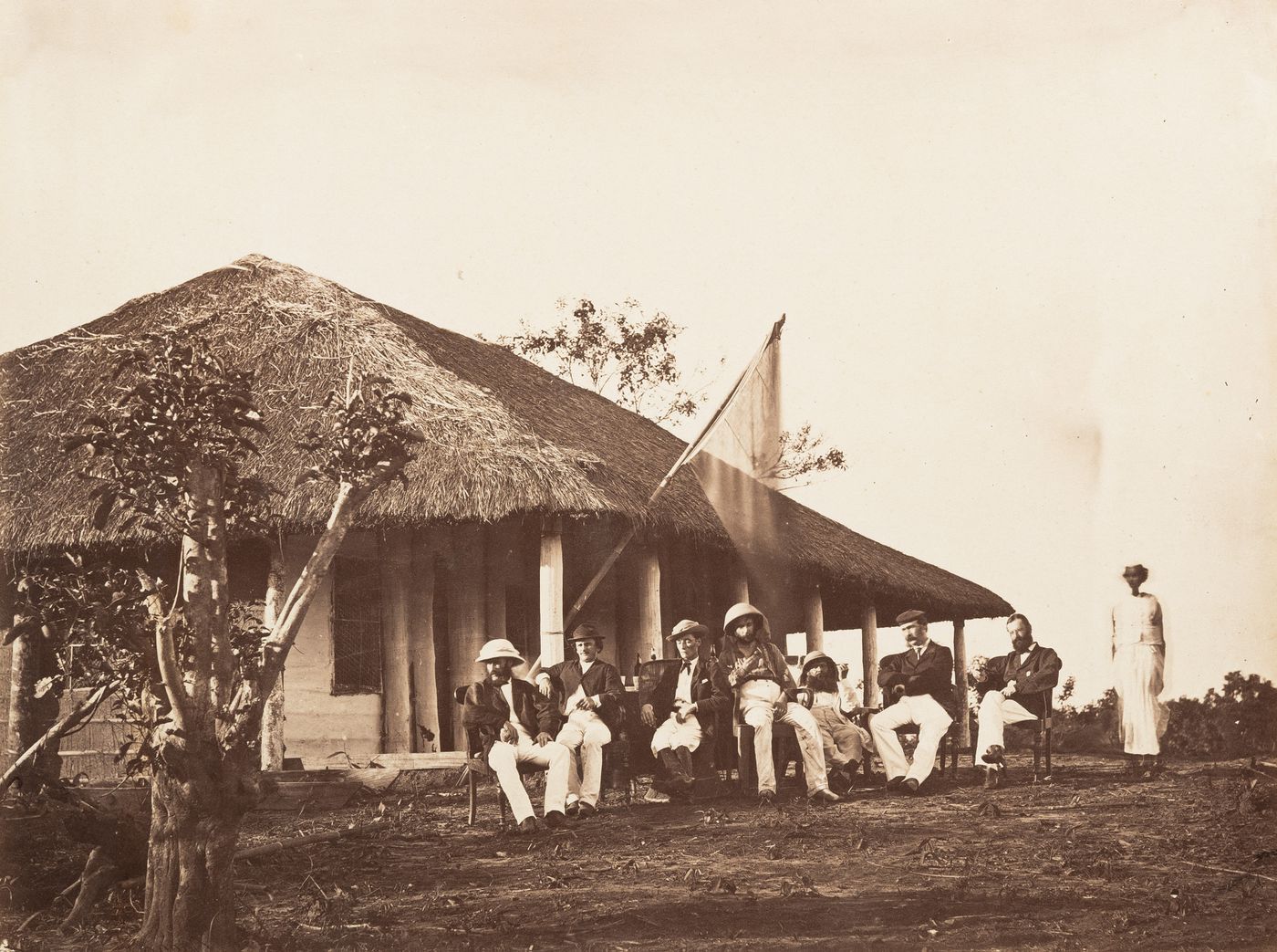Group portrait of seven tea planters sitting and woman standing before thatched bungalow, probably Assam, India